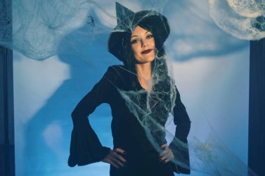 A charming witch poses in front of spider webs, her hands raised in a playful gesture. She wears a pointed hat and a confident smile, creating a whimsical Halloween atmosphere clipart