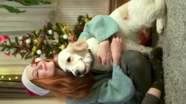 Noel Baba şapkası takan genç bir kadın rahat bir yastıkta altın av köpeğini kucaklıyor. Arka plandaki şenlikli Noel ağacı iç açıcı bir tatil atmosferi yaratıyor.