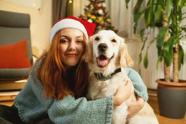 Noel Baba şapkası takan genç bir kadın arka planda güzel süslenmiş bir Noel ağacı ile yerde otururken Golden Retriever 'ına sevgiyle sarılıyor.