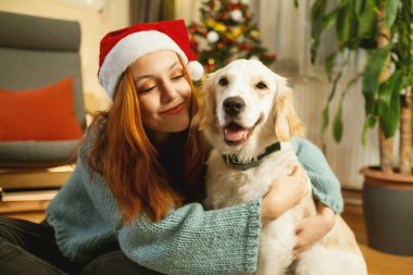 Noel Baba şapkası takan genç bir kadın arka planda güzel süslenmiş bir Noel ağacı ile yerde otururken Golden Retriever 'ına sevgiyle sarılıyor.