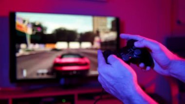 Yarış video oyunu oynarken elinde oyun kumandası olan bir adamın yakın çekim görüntüsü. Arka planda bir araba yarışını gösteren bulanık bir TV ekranı var