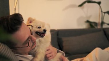 Bir erkek Pomeranian köpeğinin, sahibinin kucağında oturup uzaktan kumandayla televizyon seyretmeye çalışmasını gösteren bir video..