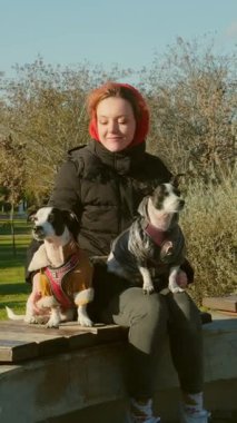 20 'li yaşlarda bir kadın parkta kucağında iki Jack Russell Teriyeri ile sonbahar güneşinin altında huzurlu bir anın tadını çıkarıyor.