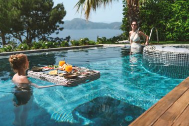 20 'li yaşlarda, havuzda dinlenirken mayo giyen sarışın ve esmer bir kadın yüzen bir kahvaltıdan zevk alır..