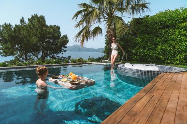 20 'li yaşlarda, havuzda dinlenirken mayo giyen sarışın ve esmer bir kadın yüzen bir kahvaltıdan zevk alır..