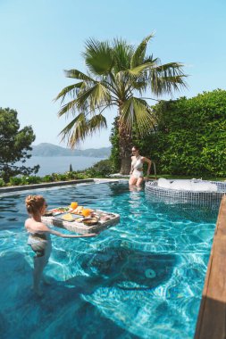 20 'li yaşlarda, havuzda dinlenirken mayo giyen sarışın ve esmer bir kadın yüzen bir kahvaltıdan zevk alır..