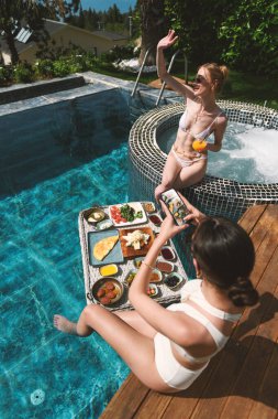 Sarışın ve esmer, 20 'li yaşlarda mayo giyip havuz kenarında dinlenirken suda yüzen kahvaltının tadını çıkarıyor.