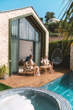 20 'li yaşlarda, rahat elbiseler giyen sarışın ve esmer biri özel havuzlarının kenarında oturup taze meyvelerin ve lüks ortamlarının tadını çıkarıyor.