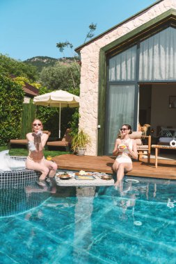 Sarışın ve esmer, 20 'li yaşlarda mayo giyip havuz kenarında dinlenirken suda yüzen kahvaltının tadını çıkarıyor.