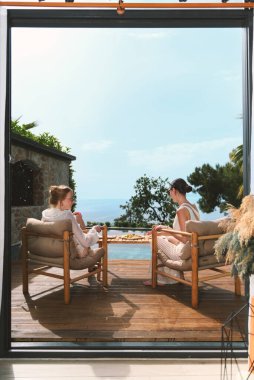 20 'li yaşlarda sarışın ve esmer bir kadın, özel havuzlarının yanında otururken rahat elbiseler giyer, taze meyvelerin ve çarpıcı deniz manzarasının tadını çıkarır..