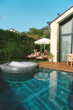 20 'li yaşlarda iki kadın, bir sarışın ve bikinili bir esmer, yemyeşil havuz kenarında güneşlenirler, kokteyllerini yudumlarken manzaranın ve güneşin tadını çıkarırlar..