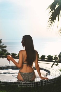 20 'li yaşlarda, bikinili esmer bir kadın özel havuzunda ve köpüklü jakuzisinde oturmuş manzaranın tadını çıkarırken elinde kokteyl tutuyor..