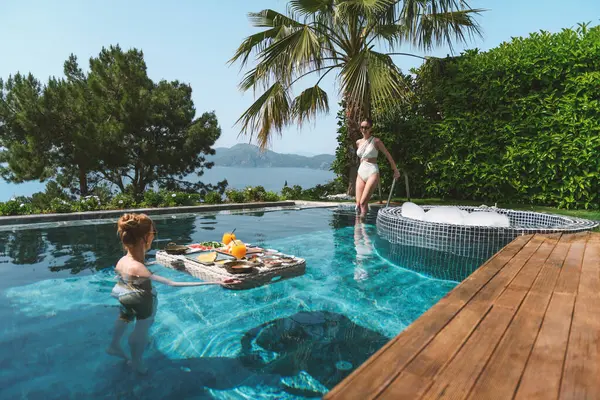 20 'li yaşlarda, havuzda dinlenirken mayo giyen sarışın ve esmer bir kadın yüzen bir kahvaltıdan zevk alır..