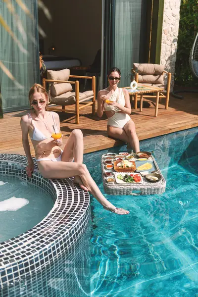 Sarışın ve esmer, 20 'li yaşlarda mayo giyip havuz kenarında dinlenirken suda yüzen kahvaltının tadını çıkarıyor.