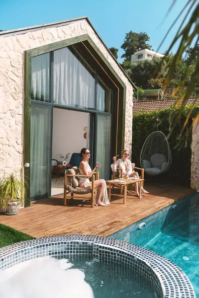 20 'li yaşlarda, rahat elbiseler giyen sarışın ve esmer biri özel havuzlarının kenarında oturup taze meyvelerin ve lüks ortamlarının tadını çıkarıyor.