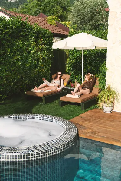 20 'li yaşlarda iki kadın, bir sarışın ve bikinili bir esmer, yemyeşil havuz kenarında güneşlenirler, kokteyllerini yudumlarken manzaranın ve güneşin tadını çıkarırlar..