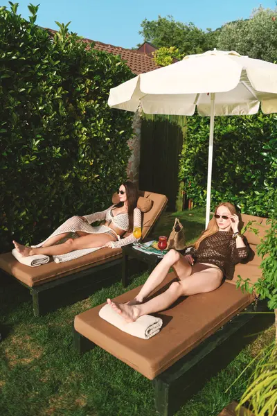 20 'li yaşlarda iki kadın, bir sarışın ve bikinili bir esmer, yemyeşil havuz kenarında güneşlenirler, kokteyllerini yudumlarken manzaranın ve güneşin tadını çıkarırlar..