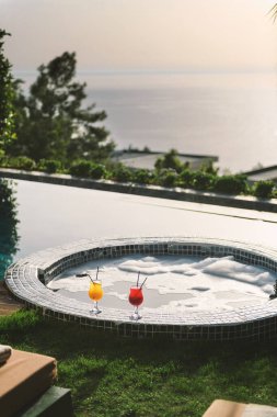 Havuz başında lüks bir otel odasında, baloncuklu jakuzinin yanına yerleştirilmiş iki canlı kokteyl ya da meyve içeceği..