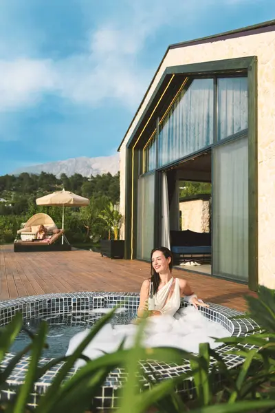 20 'li yaşlarda esmer bir kadın özel spa' sının köpüklü sularında dinleniyor, gün ışığında güneşlenirken bir yudum içki içiyor..