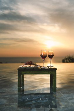 İki kadeh kırmızı şarap ve gün batımında sonsuz bir havuz tarafından sunulan zarif bir meyve tabağı..