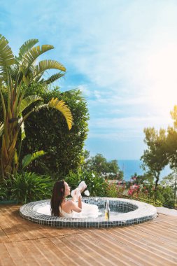 20 'li yaşlarda esmer bir kadın özel spa' sının köpüklü sularında dinleniyor, gün ışığında güneşlenirken bir yudum içki içiyor..