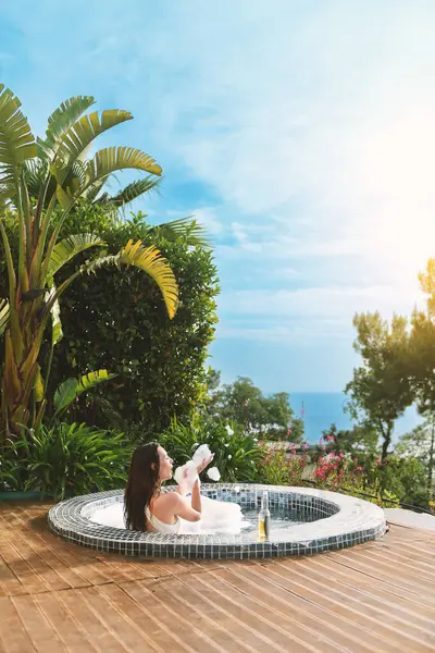 20 'li yaşlarda esmer bir kadın özel spa' sının köpüklü sularında dinleniyor, gün ışığında güneşlenirken bir yudum içki içiyor..