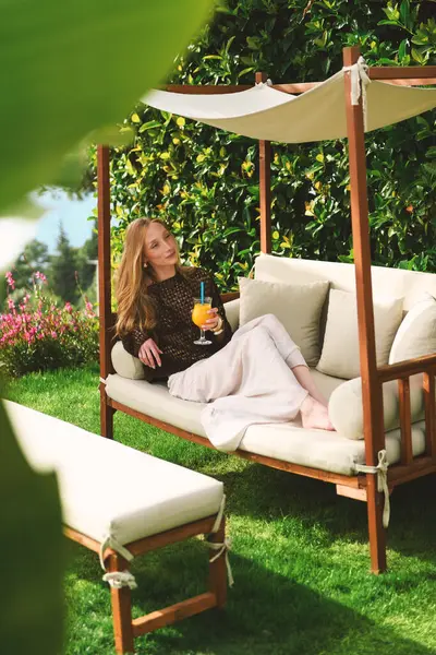 20 'li yaşlarda sarışın bir kadın bahçe sandalyesinde oturmuş portakal suyu yudumluyor ve otelinin özel bahçesindeki kameraya gülümsüyor..