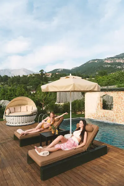 20 'li yaşlarda bir esmer ve sarışın özel havuzlarının yanında güneşli yataklarda dinleniyor ve birlikte ferahlatıcı içeceklerin keyfini çıkarıyorlar..