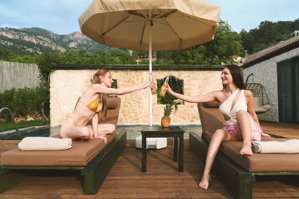 20 'li yaşlarda bir esmer ve sarışın özel havuzlarının yanında güneşli yataklarda dinleniyor ve birlikte ferahlatıcı içeceklerin keyfini çıkarıyorlar..