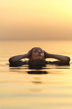 20 'li yaşlarda sarışın bir kadın gün batımında sonsuz bir havuzda yüzer. Kafası suyun üstünde, saçlarını zarifçe silerken..