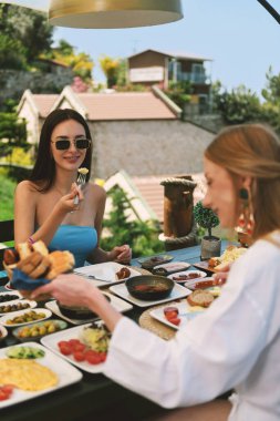 20 'li yaşlarda bir esmer ve sarışın güzel Akdeniz manzaralı bir otel masasında kahvaltı yapıyorlar..