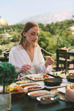 20 'li yaşlarda sarışın bir kadın, arka planda çarpıcı bir dağ manzarası olan bir otel masasında kahvaltının tadını çıkarıyor..