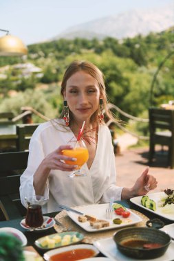 20 'li yaşlarda sarışın bir kadın, arka planda çarpıcı bir dağ manzarası olan bir otel masasında kahvaltının tadını çıkarıyor..