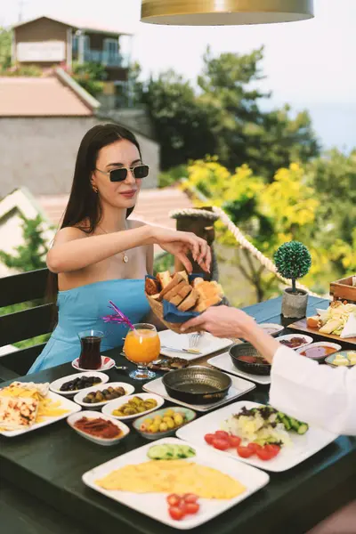 20 'li yaşlarda esmer bir kadın, arka planda çarpıcı Akdeniz manzaralı bir otel masasında kahvaltısının tadını çıkarıyor..