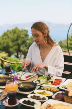 20 'li yaşlarda sarışın bir kadın, arka planda nefes kesici deniz manzarası olan bir otel masasında kahvaltının tadını çıkarıyor..