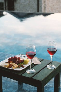İki kırmızı şarap kadehi ve havuz kenarında güzel hazırlanmış bir meyve tabağı..