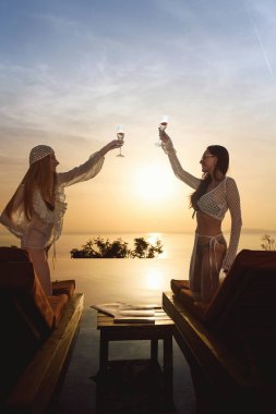 20 'li yaşlarda esmer ve sarışın bir kadın havuz kenarında gün batımında güneşli yataklarda şampanya bardakları tokuşturur..