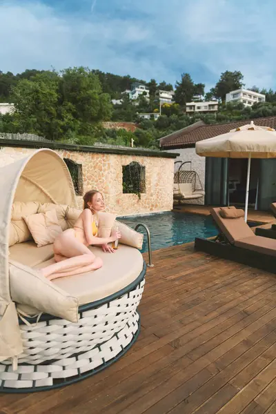 20 'li yaşlarda sarışın bir kadın özel havuzunun yanında yuvarlak bir gündüz yatağında dinleniyor. Güneş ışığı ve serinletici bir içeceğin tadını çıkarıyor..