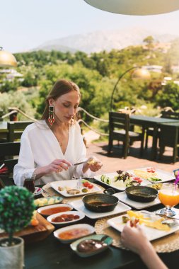 20 'li yaşlarda sarışın bir kadın, arka planda çarpıcı bir dağ manzarası olan bir otel masasında kahvaltının tadını çıkarıyor..
