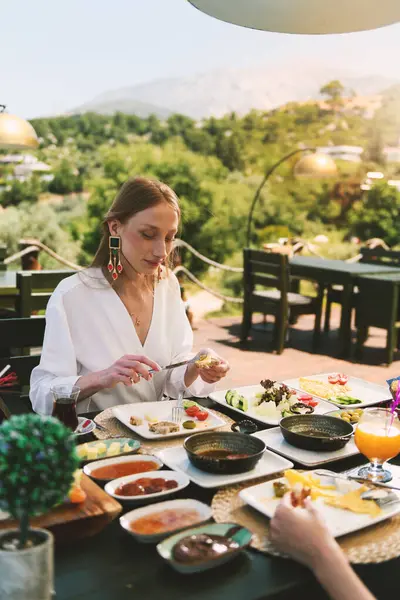 20 'li yaşlarda sarışın bir kadın, arka planda çarpıcı bir dağ manzarası olan bir otel masasında kahvaltının tadını çıkarıyor..