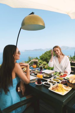 20 'li yaşlarda bir esmer ve sarışın güzel Akdeniz manzaralı bir otel masasında kahvaltı yapıyorlar..