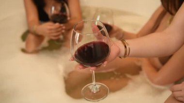 Köpüklü bir küvette dinlenen üç genç kadın. Biri yavaş çekimde kıvrılıp kırmızı şarabı yudumluyor.