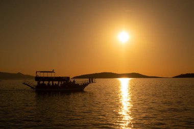 Canlı bir günbatımına karşı turist teknesinin çarpıcı bir silueti, denizin ve gökyüzünün dingin güzelliğini ılık renklerde yakalıyor.