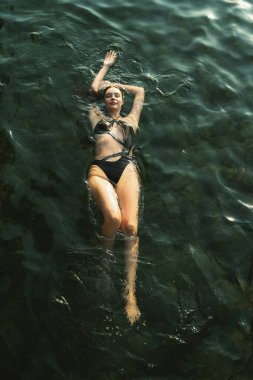 Okyanus sularında dinlenen esmer bir kadının altın gün batımı ışığı altında siyah bikini giydiği göz kamaştırıcı bir kuş bakışı fotoğrafı..