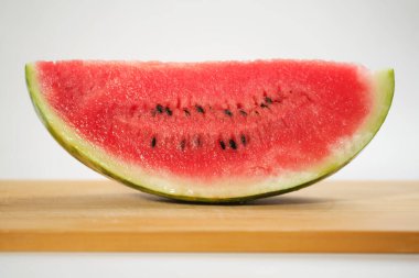 Tahta bir tahtadaki karpuz diliminin, sulu dokusunu ve canlı kırmızı rengini gözler önüne seren, beyaz bir zemine yakın plan fotoğrafı.