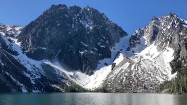 The stunning view of snow-capped Dragontail and Colchuck Peaks and Asgaard Pass towering above the still waters of alpine Colchuck Lake. Taken on a sunny blue sky day - Nr Leavenworth, Washington, USA