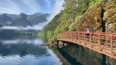 A female hiker crosses a red metal bridge over a still lake surrounded by forest and with views of mountains and clouds. Known as the Devils Punchbowl - Lake Crescent, Washington, USA