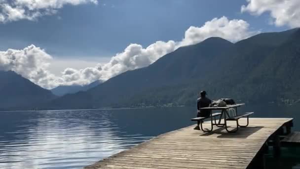 美国华盛顿州新月湖 一位女徒步旅行者坐在木制码头上的野餐长椅上 俯瞰着美丽的波涛汹涌的湖面 背景是森林覆盖的群山和云彩 — 图库视频影像