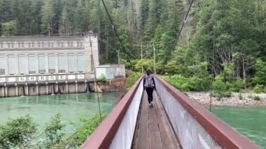 Kuzey Cascades 'teki Skagit Nehri üzerindeki asma köprüden geçen bir kadın. Vadi hidroelektrik santrali ve her zaman yeşil olan ormanlar arka planda, Newhalem, Washington, ABD
