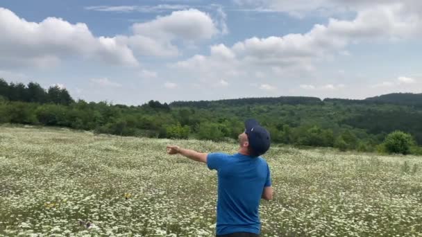 青いTシャツを着た男が 背景に森のある美しい白い野の花の草原の真ん中にドローンを手に入れます ブルガリア ヴェリコ タルノヴォ氏 — ストック動画